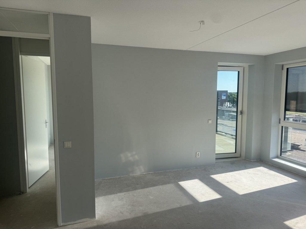 Lichte nieuwbouwkamer met lichtgrijze wanden, een betonvloer, een balkondeur en zicht op een bouwterrein