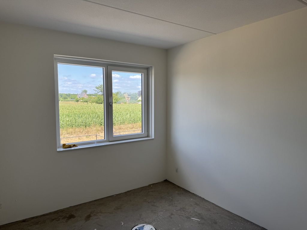Slaapkamer in een nieuwbouwwoning met strak afgewerkte witte wanden en een raam dat uitkijkt op een maisveld