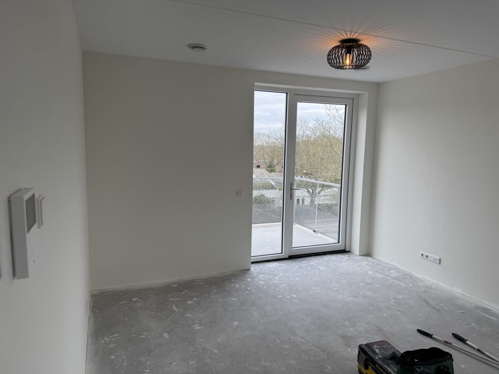 Nieuwe kamer met egaal witte wanden, een balkondeur en een kale dekvloer, klaar voor de vloerlegger