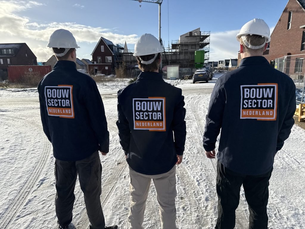 Drie vakmensen van Bouwsector Nederland met witte bouwhelm en donkere jas, van achteren gezien, op een besneeuwd bouwterrein met huizen in aanbouw